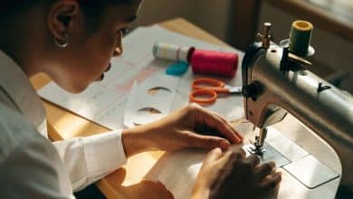 Pessoa aprendendo a costurar em uma máquina doméstica, com tecido, linhas coloridas, tesoura e moldes organizados sobre uma mesa iluminada.