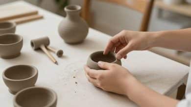 Mãos de uma pessoa iniciante modelando uma pequena tigela de argila sobre uma bancada clara, com rolinho, ferramentas simples e texturas naturais ao fundo, em um ateliê acolhedor.
